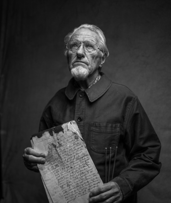 Zwart-wit portret van een man met bril die een oud, versleten document vasthoudt. (vergroot foto)