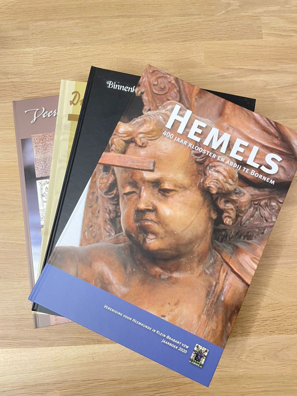 Vier boeken liggen op een houten tafel, het bovenste boek heeft een afbeelding van een houten beeldje en de titel "HEMELS". (vergroot foto)