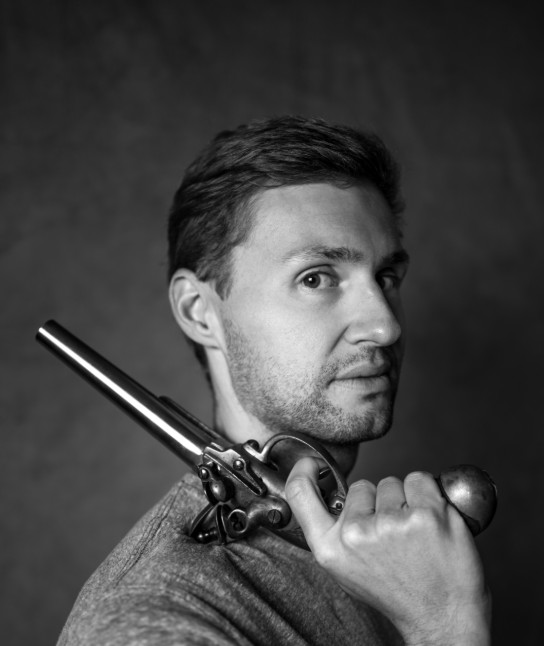 Zwart-wit portret van een man die een oude revolver op zijn schouder houdt en naar de camera kijkt. (vergroot foto)