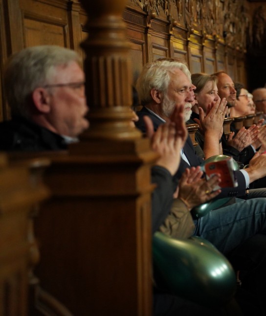 Mensen applaudisseren zittend in een zaal met een scherm op de achtergrond. (vergroot foto)