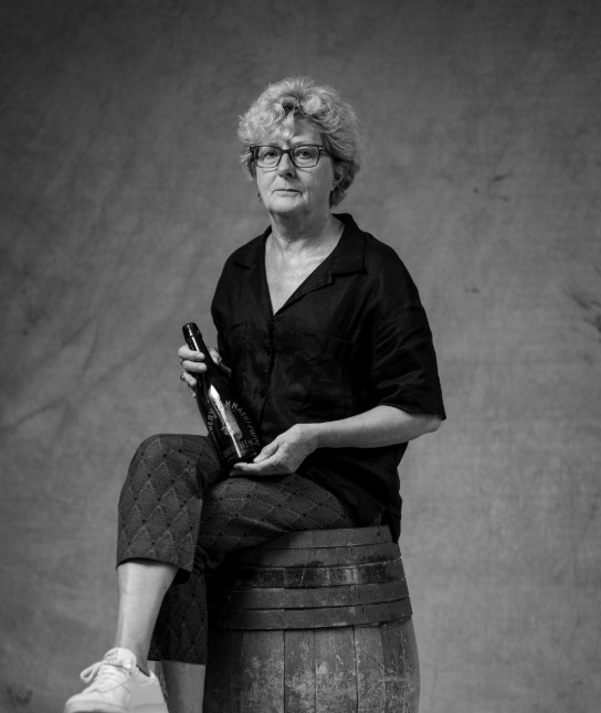 Zwart-wit portret van een vrouw met bril, zittend op een houten vat, met een fles in haar handen. (vergroot foto)